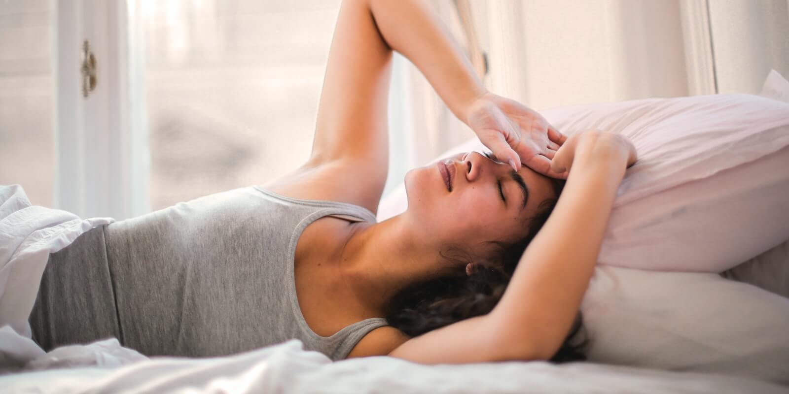 Woman in grey tank top lying in bed having headache and body pain